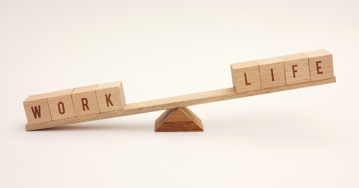 Wooden alphabet blocks a top a long wooden board teetering on a wooden fulcrum implying a teeter-totter. The left side blocks spell 'WORK' and are weighing down it's side of the teeter-totter with respect to the other side's blocks which spell 'LIFE', implying the two sides of work and life are out of balance