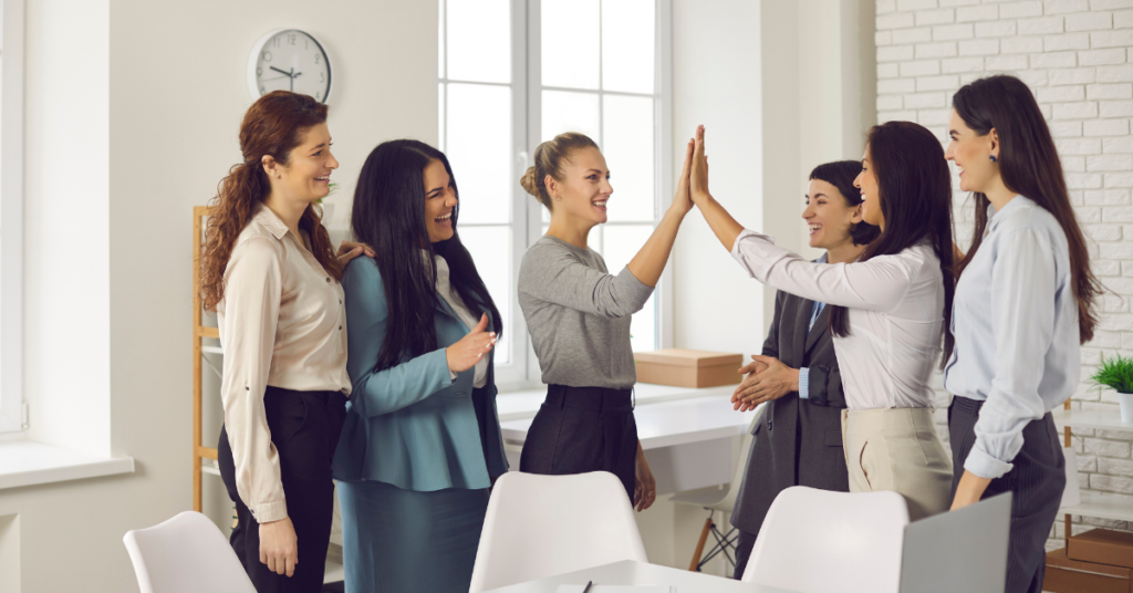 Women smiling and standing around a desk looking at each other and holding hands up for a high five